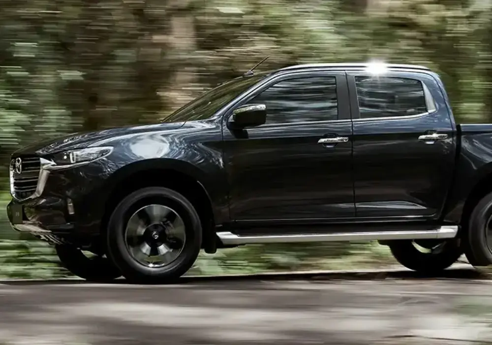 Mazda BT-50 negra de doble cabina conduciendo por un camino rural arbolado.